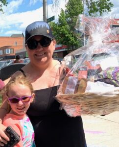 Woman in a black sundress wearing a baseball cap holding a prize basket with her arm around a child. 
